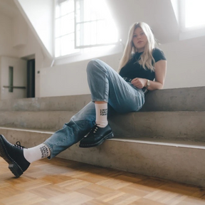 Person sitzt lässig auf einer Treppe und trägt die Statementsocken LIBERTÉ ÉGALITÉ FUCK AFD von arrel in weiß am Fuß